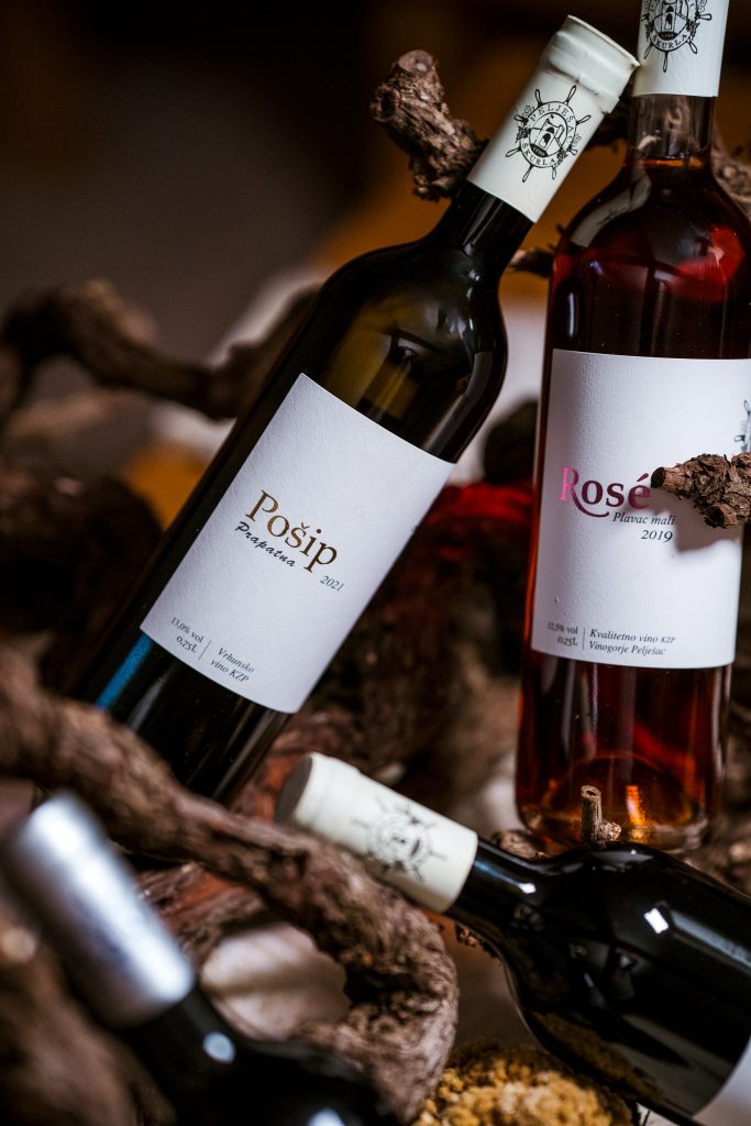Vrhunsko suho bijelo vino autohtone sorte Pošip vinarije Škurla na Pelješcu. Boca položena na trs vinove loze u rustikalnom ambijentu. S desne strane vidi se rose vino plavac mali
