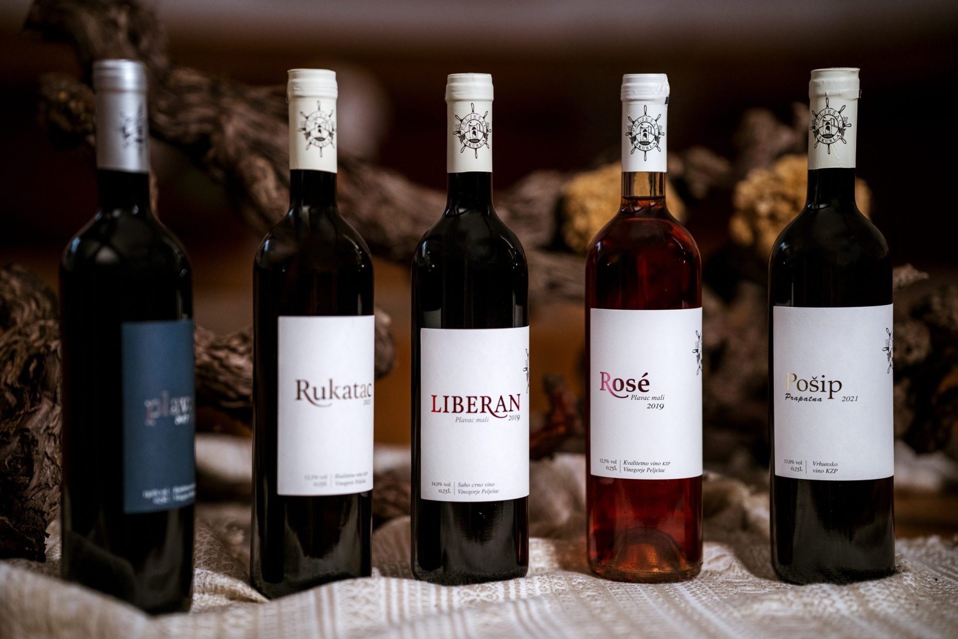 Boce vina Vinarije Škurla od autohtonih sorti poluotoka Pelješca i južne Dalmacije; kvalitetno crno suho vino plavac mali, vrhunsko vino plavac mali Liberan, kvalitetno bijelo suho vino rukatac i vrhunsko bijelo suho vino pošip, prikazane u rustikalnom ambijentu