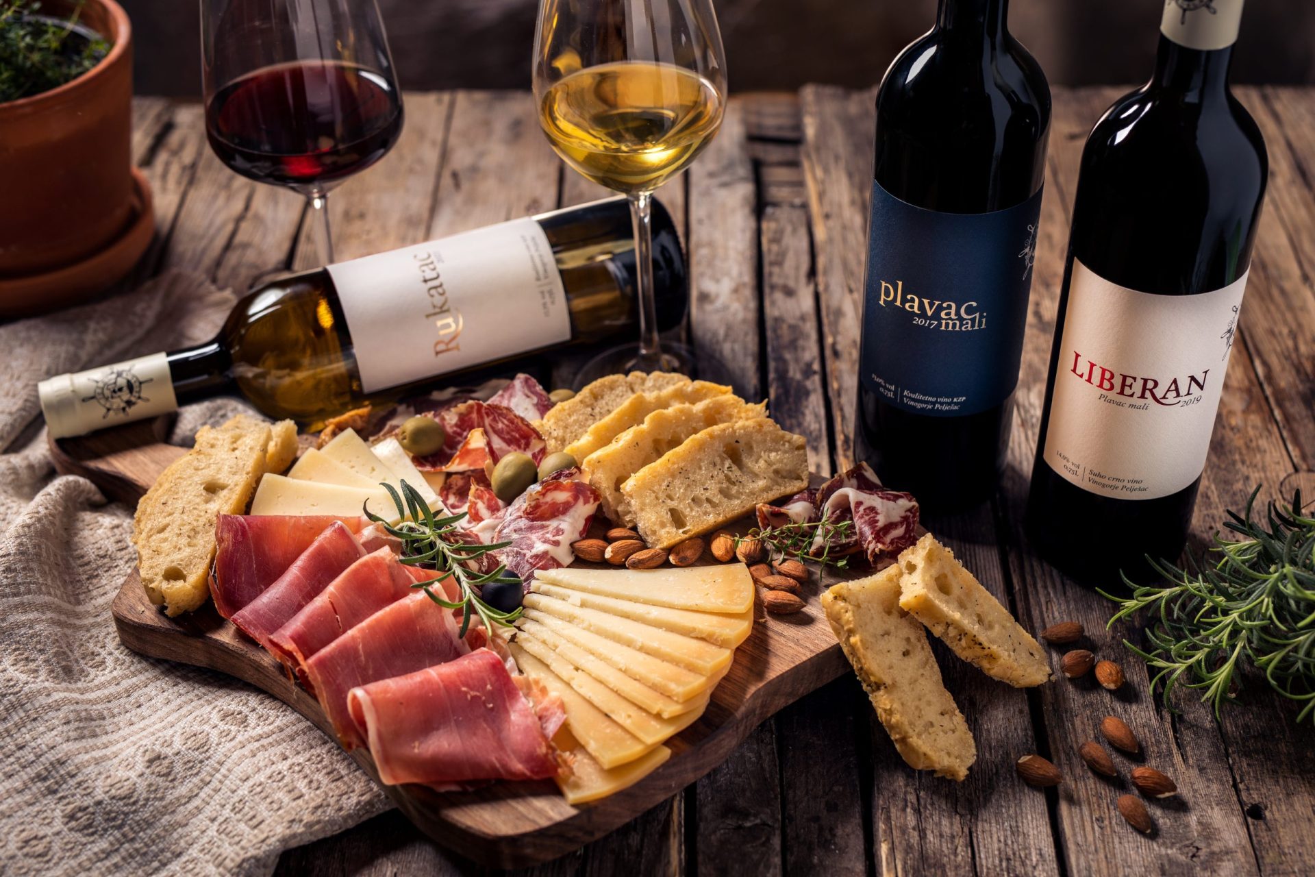 Plata s tradicionalnim dalmatinskim proizvodima: domaći kruh, kravlji i kozji sir, pršut, pečenica, masline, bademi. Vina Vinarije Škurla u bocama: vrhunsko suho crno vino plavac mali Liberan, kvalitetno suho crno vino Plavac mali i kvalitetno suho bijelo vino Rukatac. Natočene čaše crnog i bijelog vina. Prikazano na drvenom stolu u rustikalnom ambijentu