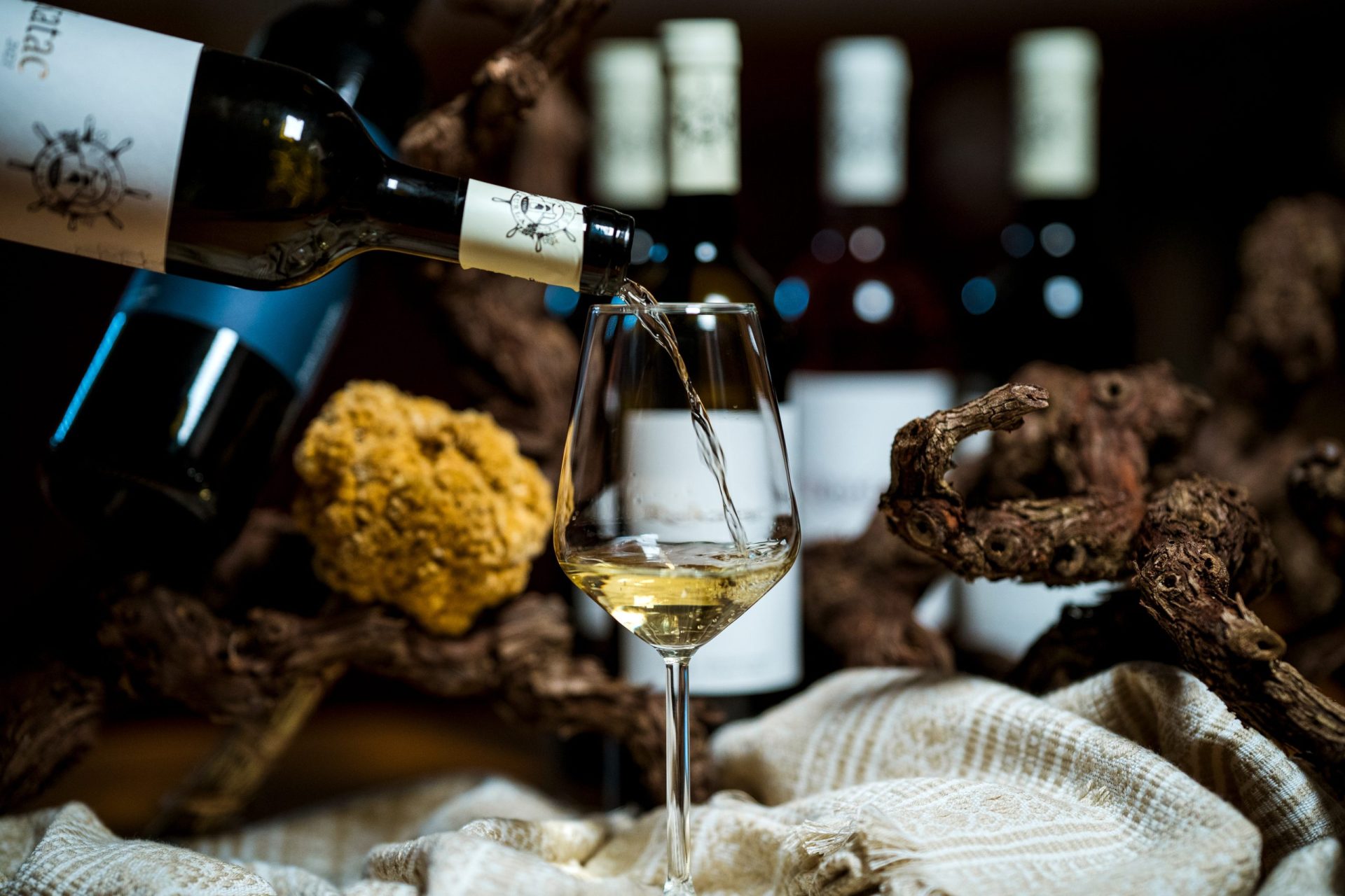 Detalj točenja bijelog vina u čašu u rustikalnom ambijentu kušaone u vinariji. na boci se nazire naziv Rukatac, u pozadini iza trsa vinove loze su vidljive druge boce vina